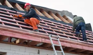 Dwaj budowlańcy pracują na dachu, montując wiatroizolację. Jeden z nich w czerwonym kasku wbija gwoździe.