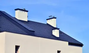 Nowy dom z ciemnym, łupkowym dachem bezokapowym i białymi kominami, pod błękitnym niebem.