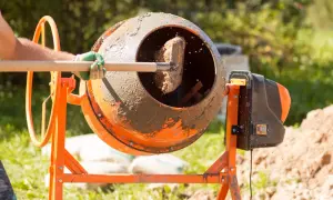 Osoba w zielonej rękawicy miesza łopatą cement w pomarańczowej betoniarce.
