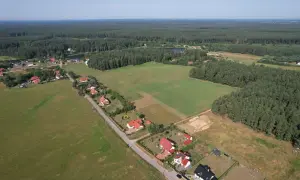 Widok z lotu ptaka na wieś otoczoną lasem. Domy z czerwonymi dachami i zielone pola. Teren ma około 1 ar.