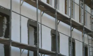 Budowa domu z bloczków styropianowych. Rusztowania otaczają białą ścianę z otworami okiennymi i drzwiami, przygotowanymi pod dalsze prace.