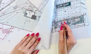 Ręka z różowymi paznokciami wskazuje ołówkiem na mapę do celów projektowych. Widoczne szczegóły działki i budynku.