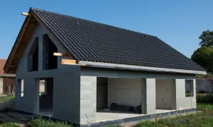 Dom w stanie surowym otwartym, z dachem pokrytym czarną dachówką, bez okien i drzwi.