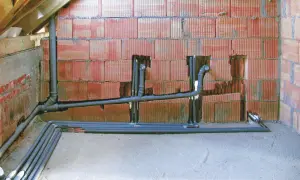 Instalacja hydrauliczna w budowie, widoczne rury i kolanka. W ścianie z czerwonej cegły przygotowane otwory na piony, kluczowe dla prawidłowego odpowietrzenia kanalizacji.