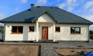 Nowoczesny dom kanadyjski z jasną elewacją, ciemnym dachem i drewnianymi oknami. Przed domem ścieżka z kostki brukowej i plac budowy.