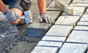 Układanie kostki brukowej na podsypce. Ręce w rękawiczkach kładą kamień, używając młotka. Widać suchy beton proporcje.