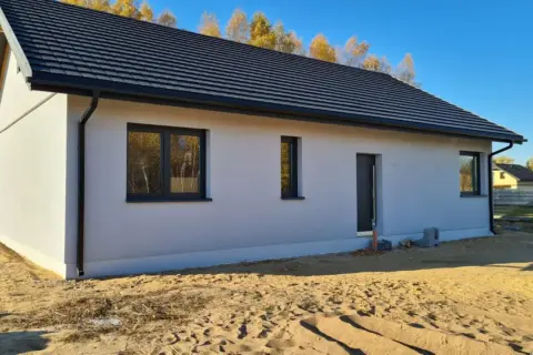 Nowy dom z szarym dachem i białymi ścianami, otoczony piaskiem i jesiennymi drzewami. Ssz.