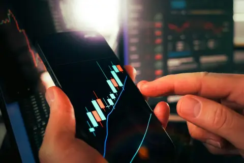 Dłoń trzyma smartfon z wykresem świecowym, pokazującym wibor 3m aktualny. W tle widać inne wykresy finansowe.