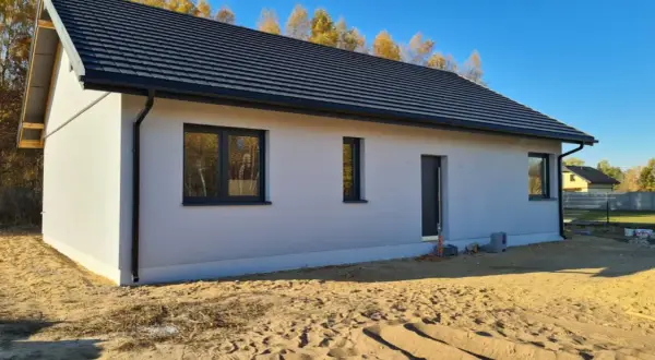 Nowy dom z szarym dachem i białymi ścianami, otoczony piaskiem i jesiennymi drzewami. Ssz.