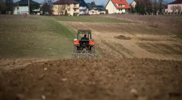 Traktor orze pole, przygotowując je pod przyszłą zabudowę. W tle widać domy, sugerujące warunki zabudowy na działce rolnej.