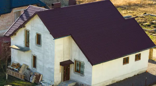 Nowy dom w zabudowie zagrodowej z ciemnobordową dachówką, w trakcie budowy.
