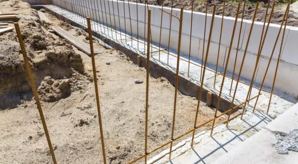 Budowa fundamentów: pionowe i poziome zbrojenie czeka na beton. W tle białe bloczki fundamentowe i usypana ziemia.