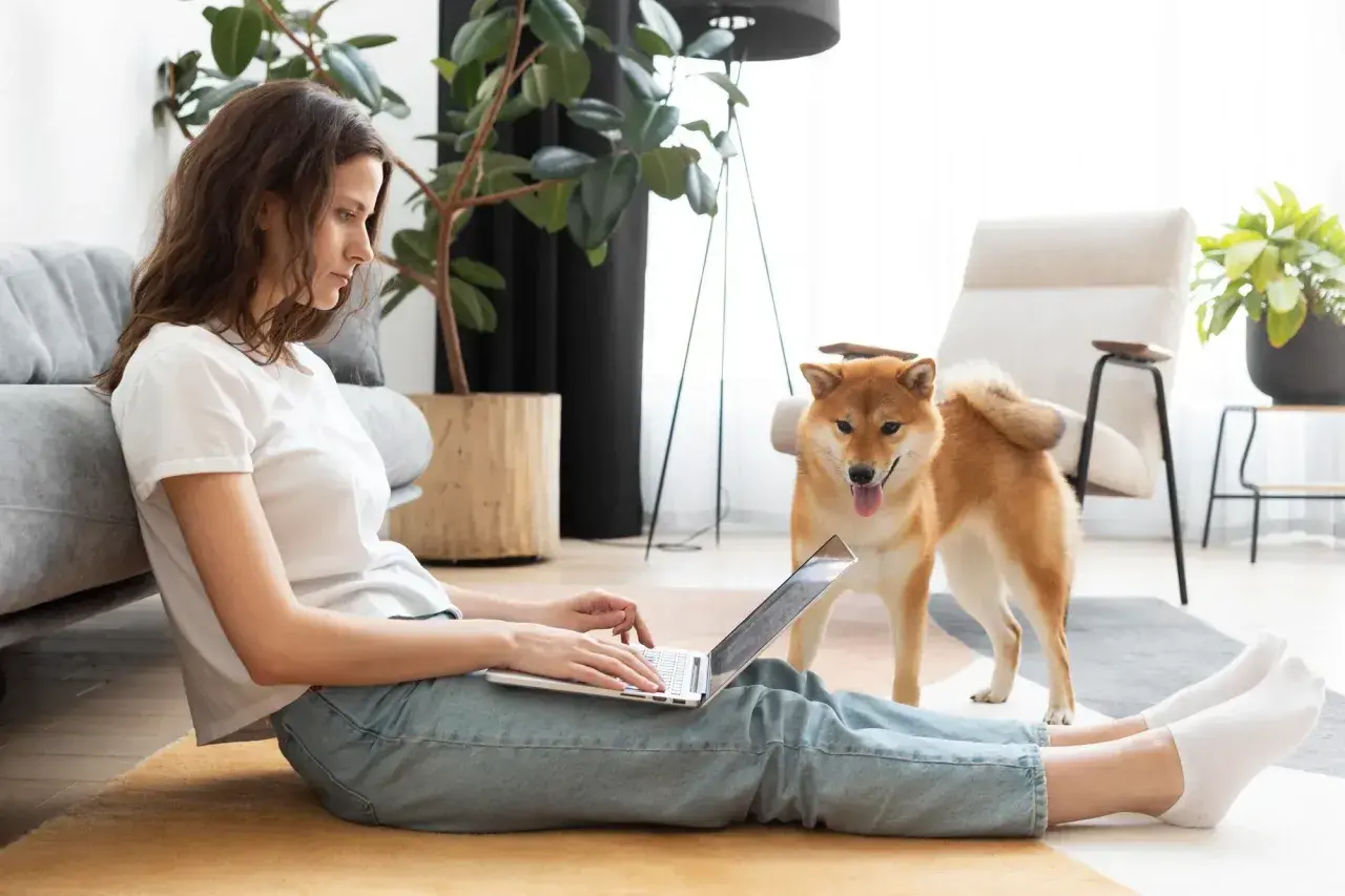 Kobieta z psem Shiba Inu pracuje na laptopie, siedząc na podłodze. Może szukać informacji o tym, jak zostać dobrym wynajmującym.