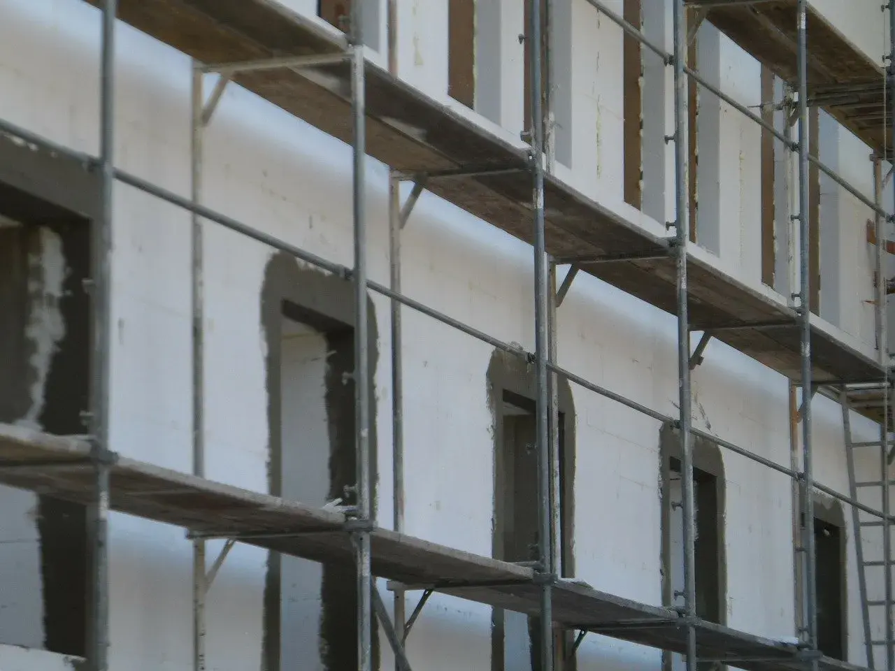 Budowa domu z bloczków styropianowych. Rusztowania otaczają białą ścianę z otworami okiennymi i drzwiami, przygotowanymi pod dalsze prace.