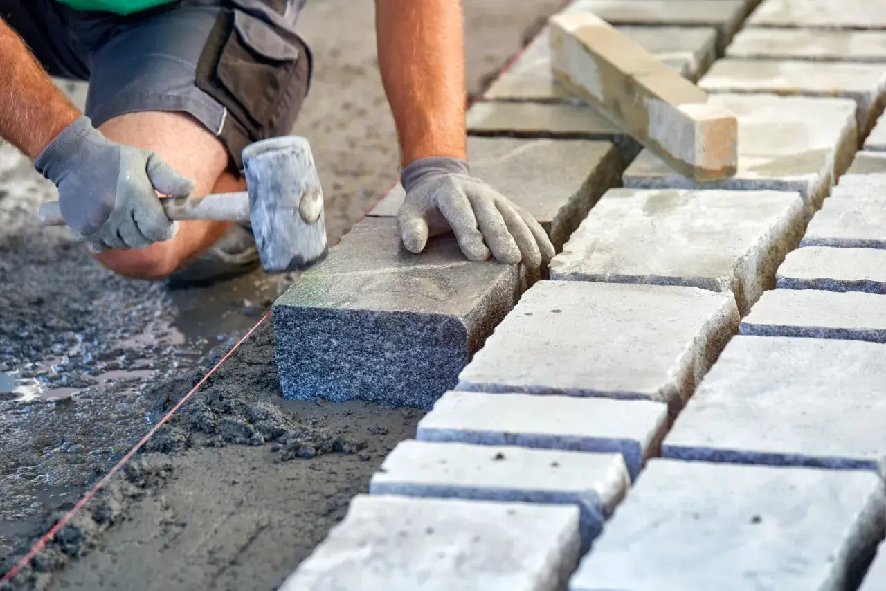 Układanie kostki brukowej na podsypce. Ręce w rękawiczkach kładą kamień, używając młotka. Widać suchy beton proporcje.
