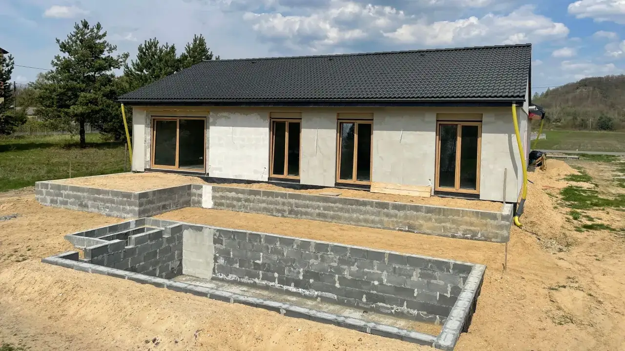 Budowa domu w toku: szary dach, betonowe fundamenty i ściany z pustaków. Wokół piasek i drzewa.