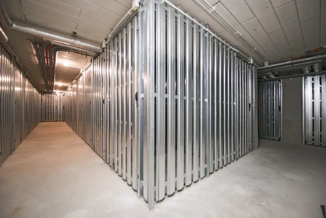 Korytarz z rzędami metalowych przegród, tworzących komórki lokatorskie. Widać instalacje biegnące pod sufitem.