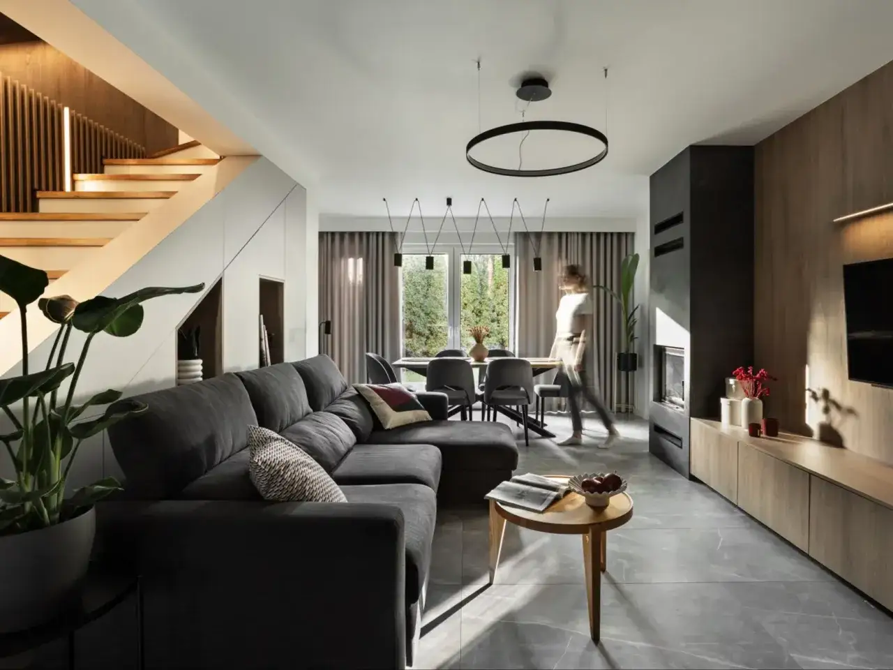 Nowoczesny salon po przebudowie z szarą kanapą, drewnianymi schodami i jadalnią.