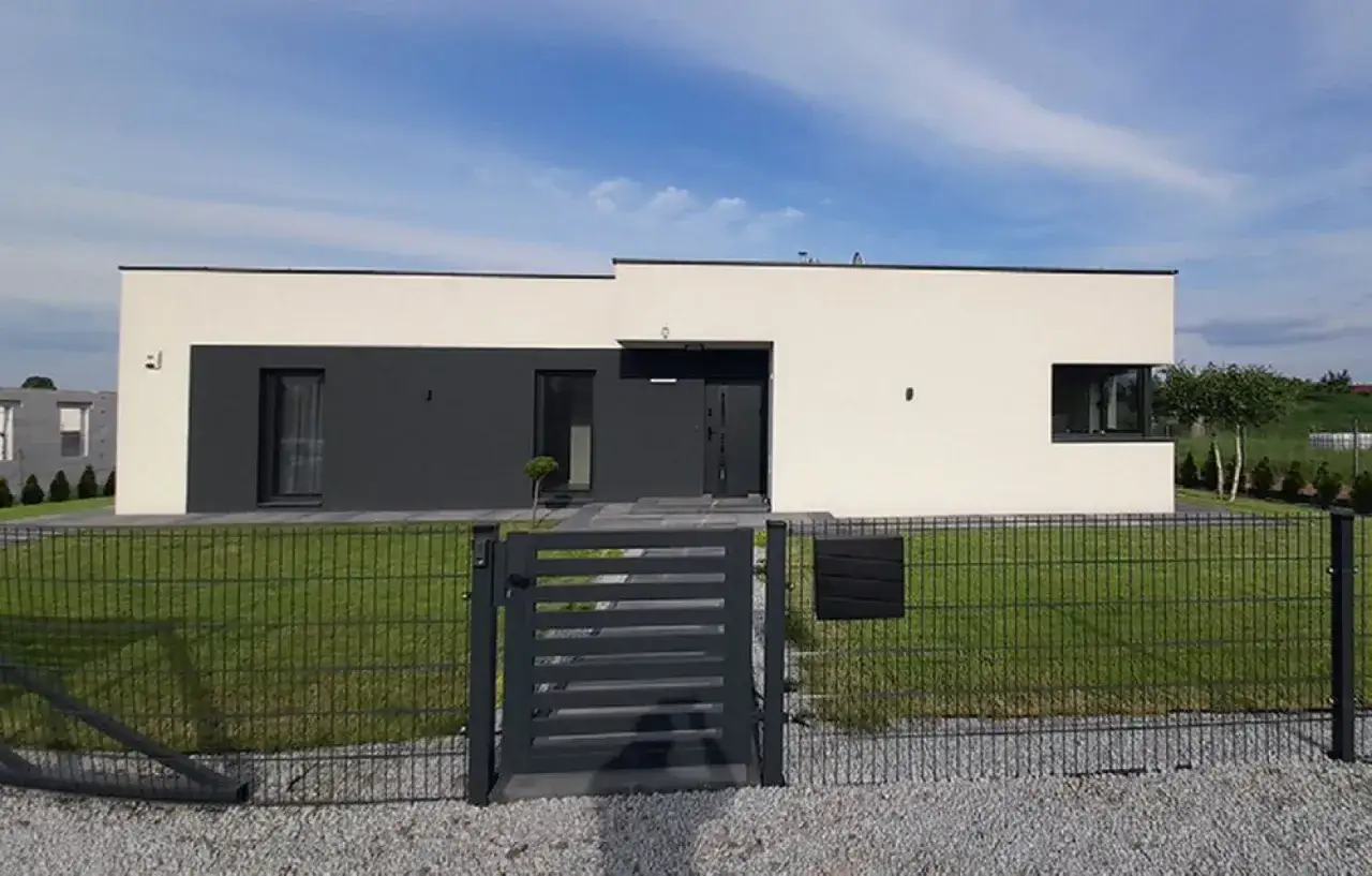 Nowoczesny dom z płaskim dachem, przypominający attikę, z dwukolorową elewacją i metalowym ogrodzeniem.