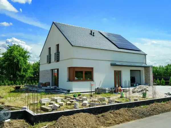 Nowy dom w trakcie budowy, z panelami słonecznymi na dachu. Rozbudowa domu postępuje, widać materiały budowlane i fundamenty.