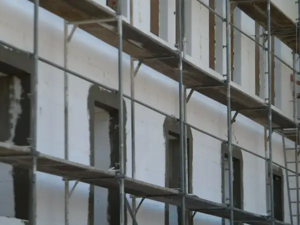 Budowa domu z bloczków styropianowych. Rusztowania otaczają białą ścianę z otworami okiennymi i drzwiami, przygotowanymi pod dalsze prace.