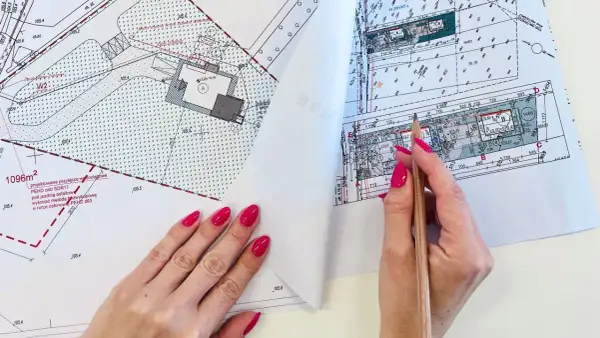 Ręka z różowymi paznokciami wskazuje ołówkiem na mapę do celów projektowych. Widoczne szczegóły działki i budynku.