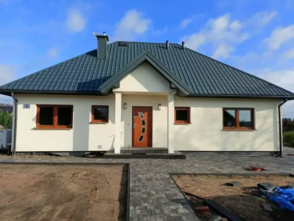 Nowoczesny dom kanadyjski z jasną elewacją, ciemnym dachem i drewnianymi oknami. Przed domem ścieżka z kostki brukowej i plac budowy.